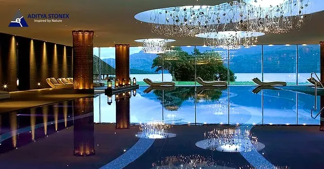 A luxury indoor pool area by Aditya Stonex featuring reflective dark flooring, statement pillars, and grand chandeliers overlooking a lake view.