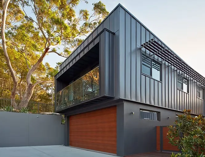 A striking modern residence showcasing dark vertical metal siding on the cantilevered upper volume, complemented by a warm wood garage door below.