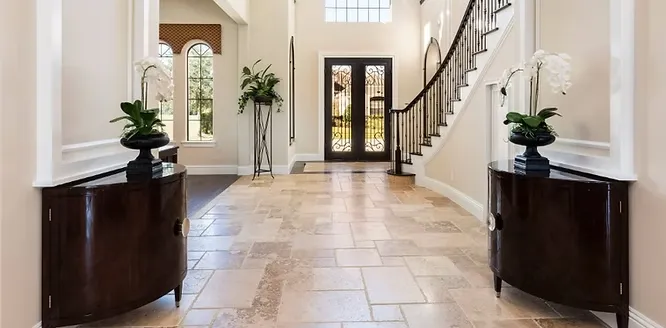 A grand home entryway featuring a tiled floor made of natural limestone in warm beige tones, leading towards a winding staircase.