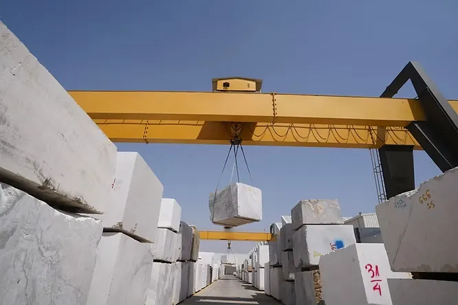 A heavy-duty yellow gantry crane hoists a massive, solid marble slab block inside a busy processing yard in Kishangarh.