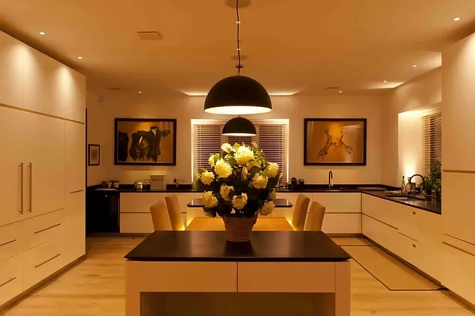 Cozy modern kitchen highlighting the effect of lighting with warm dome pendant lights over an island with white flowers.