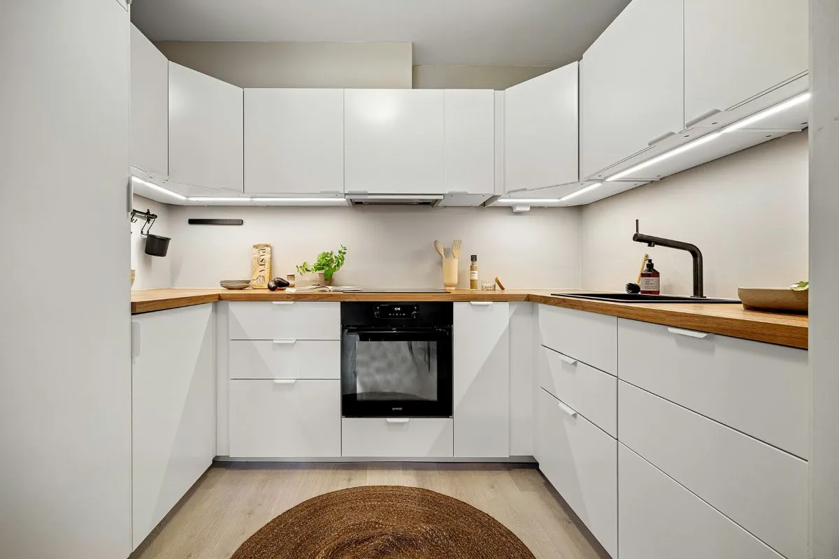A bright, U-shaped closed modular kitchen with minimalist white cabinets, wooden countertops, and an integrated oven.