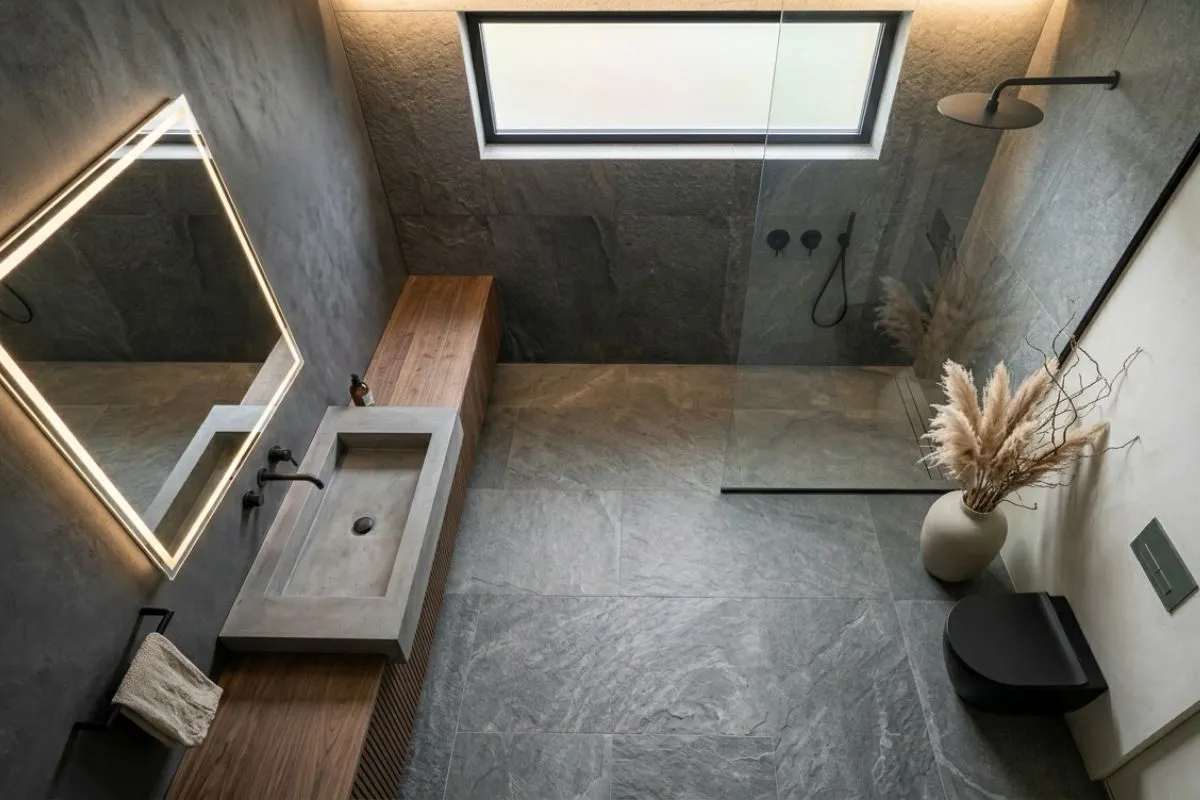 Top view of a bathroom interior showing grey stone tiles, a concrete sink, and a glass shower wall.