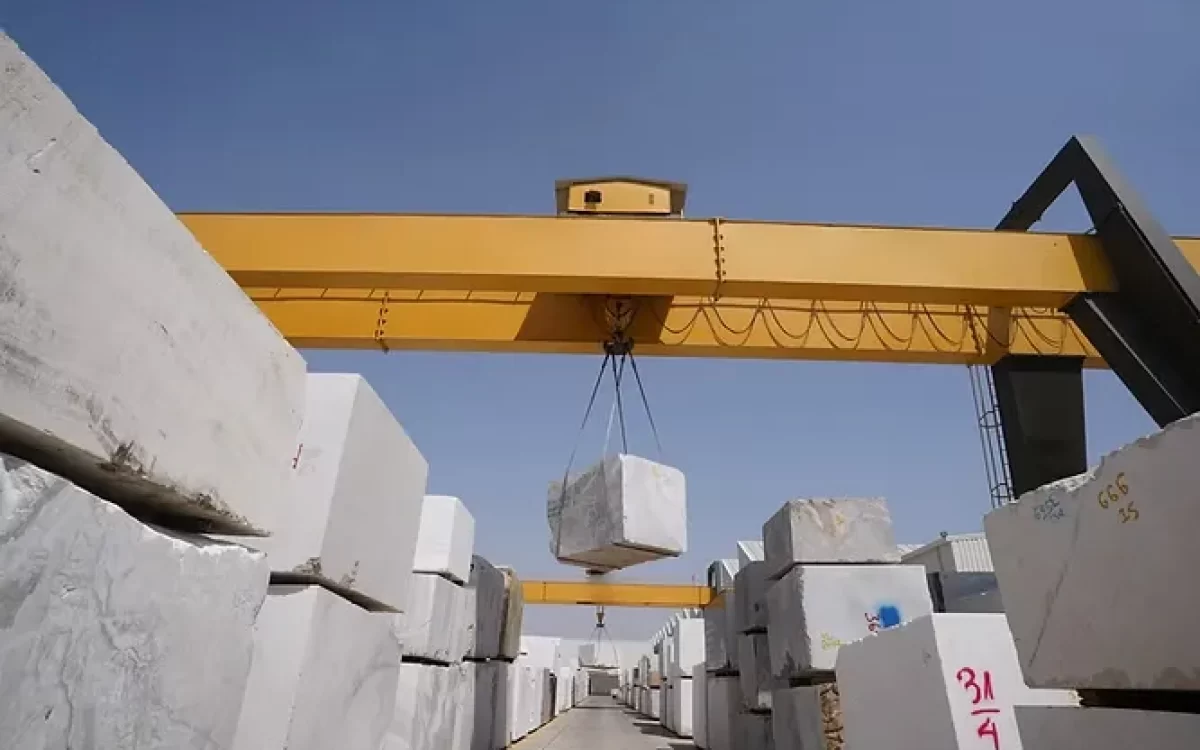 A heavy-duty yellow gantry crane hoists a massive, solid marble slab block inside a busy processing yard in Kishangarh.